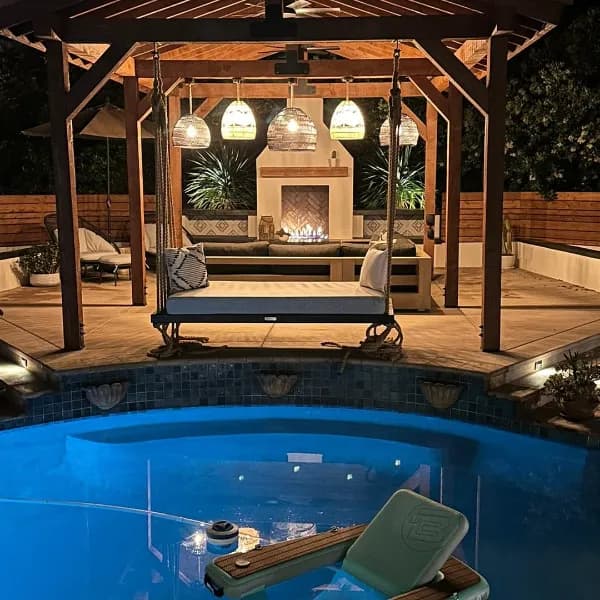 Night view of pergola-covered pool with built-in fireplace, hanging daybed swing, and spa featuring decorative tile accents and ambient lighting