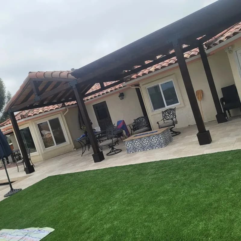 Modern aluminum patio cover extending from Mediterranean-style home with synthetic turf lawn and decorative tile fire pit table