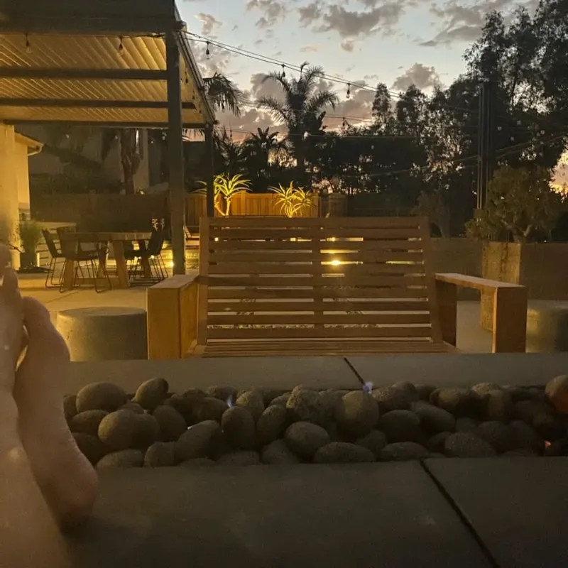 Contemporary backyard design featuring illuminated horizontal slat privacy screen, modern fire pit with river rock, and large format concrete pavers at sunset