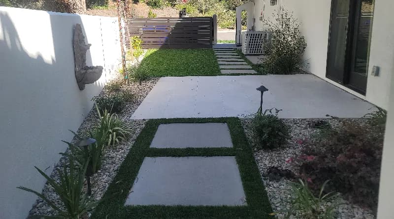 Modern backyard featuring large format concrete patio slabs with artificial turf walkway strips, decorative rock landscaping, and horizontal privacy fence in San Diego